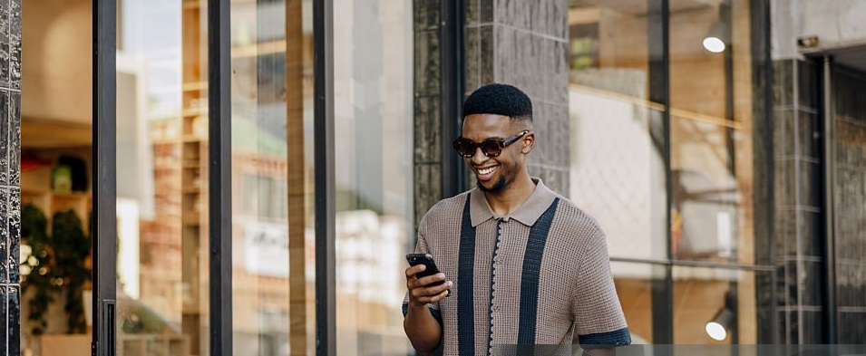 This photograph captures a stylish young Black man with a radiant smile, wearing trendy tortoiseshell sunglasses and a textured brown knit polo with a contrasting black stripe. The low-angle shot, taken outdoors against a modern architectural backdrop, enhances his confident and fashionable presence. The golden sunlight casts a warm glow, highlighting his smooth skin and well-groomed beard. His relaxed yet self-assured posture makes this image ideal for fashion, street style, urban lifestyle, and modern menswear content. Perfect for keywords like fashion photography, stylish Black man, urban street style, contemporary menswear, sunglasses trend, confident smile, and modern architecture background.