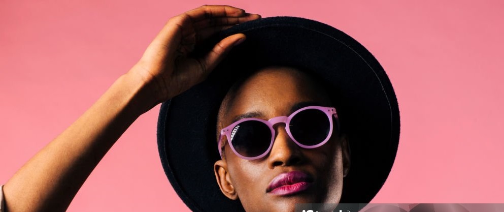 A fashion portrait of a young woman with purple sunglasses and black hat, isolated on pink studio background
